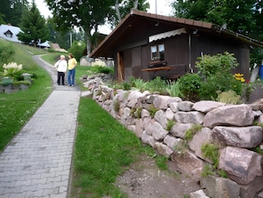 A rustic cabin with a stone wall and a wooden roof, surrounded by greenery and a paved path.