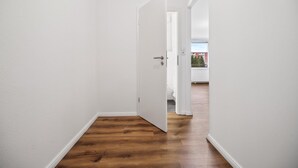 Un couloir étroit aux murs blancs et au plancher en bois, qui mène à une porte donnant sur un balcon et sur un bâtiment situé à l’extérieur.