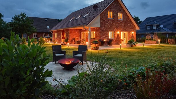 Ein Ziegelhaus mit geneigtem Dach, eine Terrasse mit Stühlen und einem Feuerkorb sowie ein gut beleuchteter Garten.