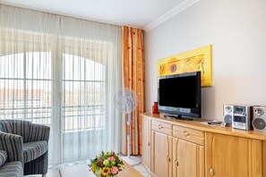 A living room with a TV, speakers, a fan, a sofa, and a window with curtains. - Apartment 'Europa' with Shared Garden, Balcony and Wi-Fi (Bad Füssing)
