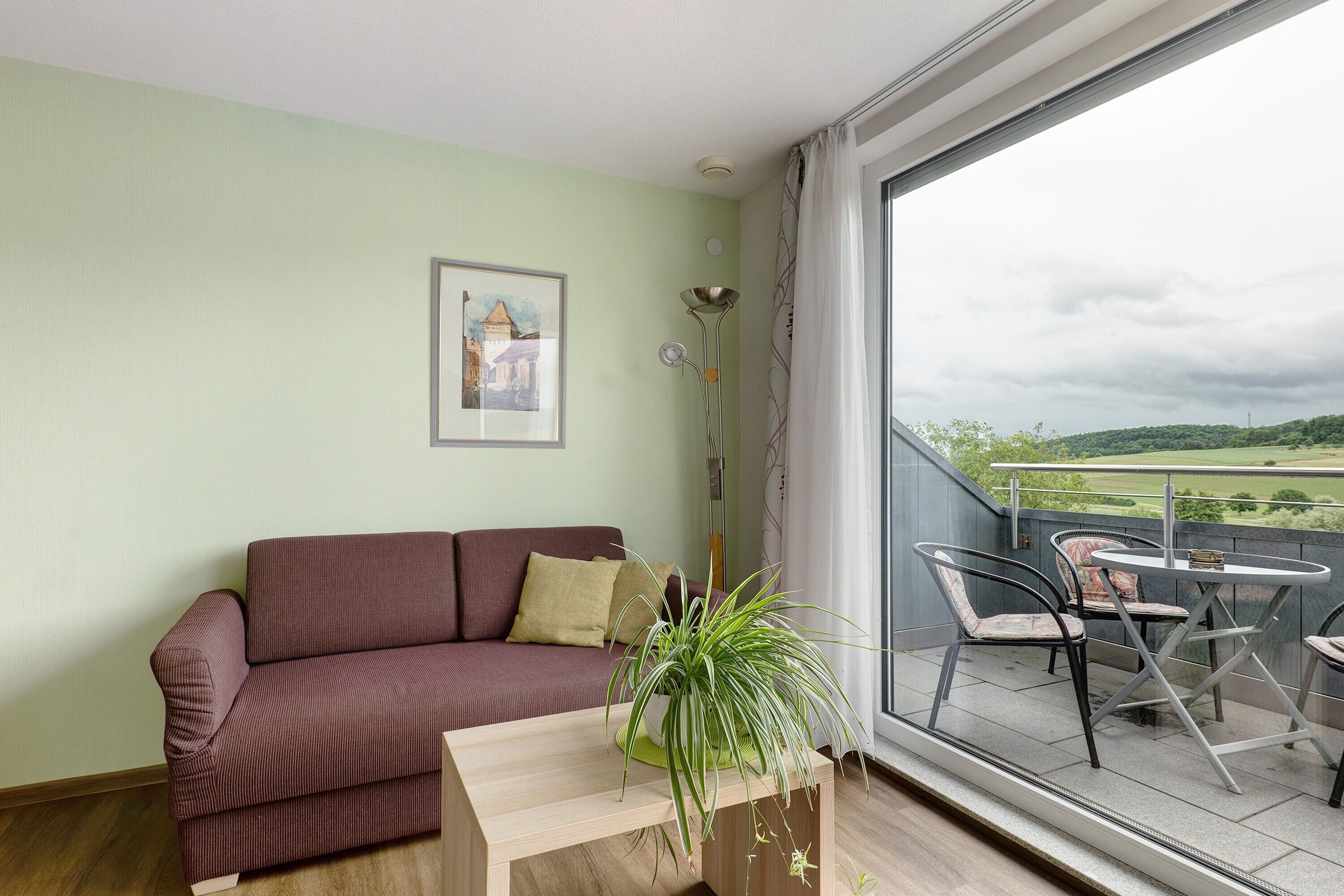 A living room with a sofa, a table, chairs, a potted plant, a picture on the wall, and a balcony with a view.
