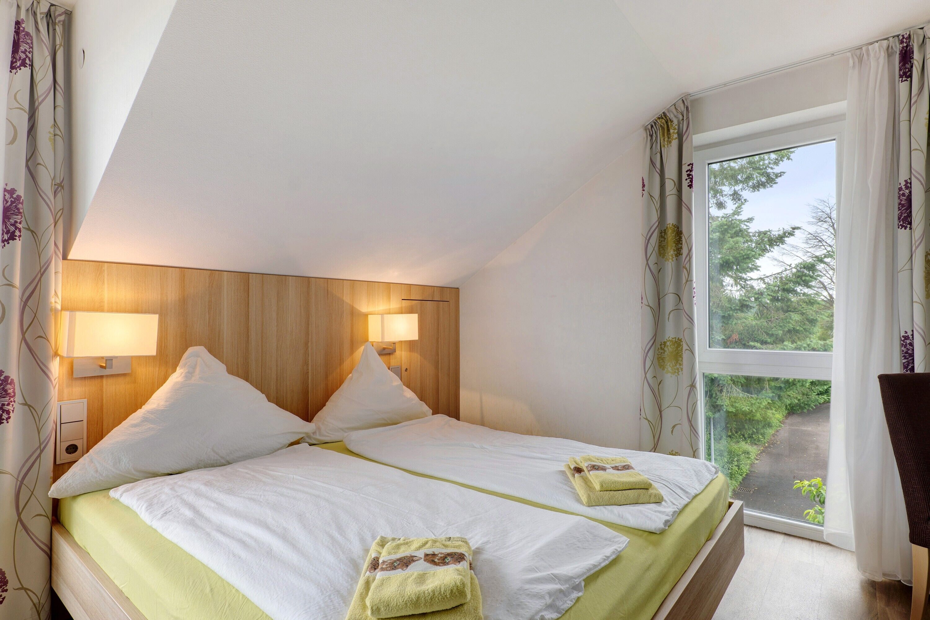 Quarto com duas camas, cabeceira de madeira, luminárias de cabeceira e vista para o verde lá fora.