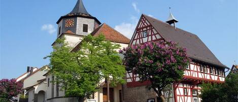 A traditional half-timbered building with a clock tower, a church, and a well-maintained garden.
