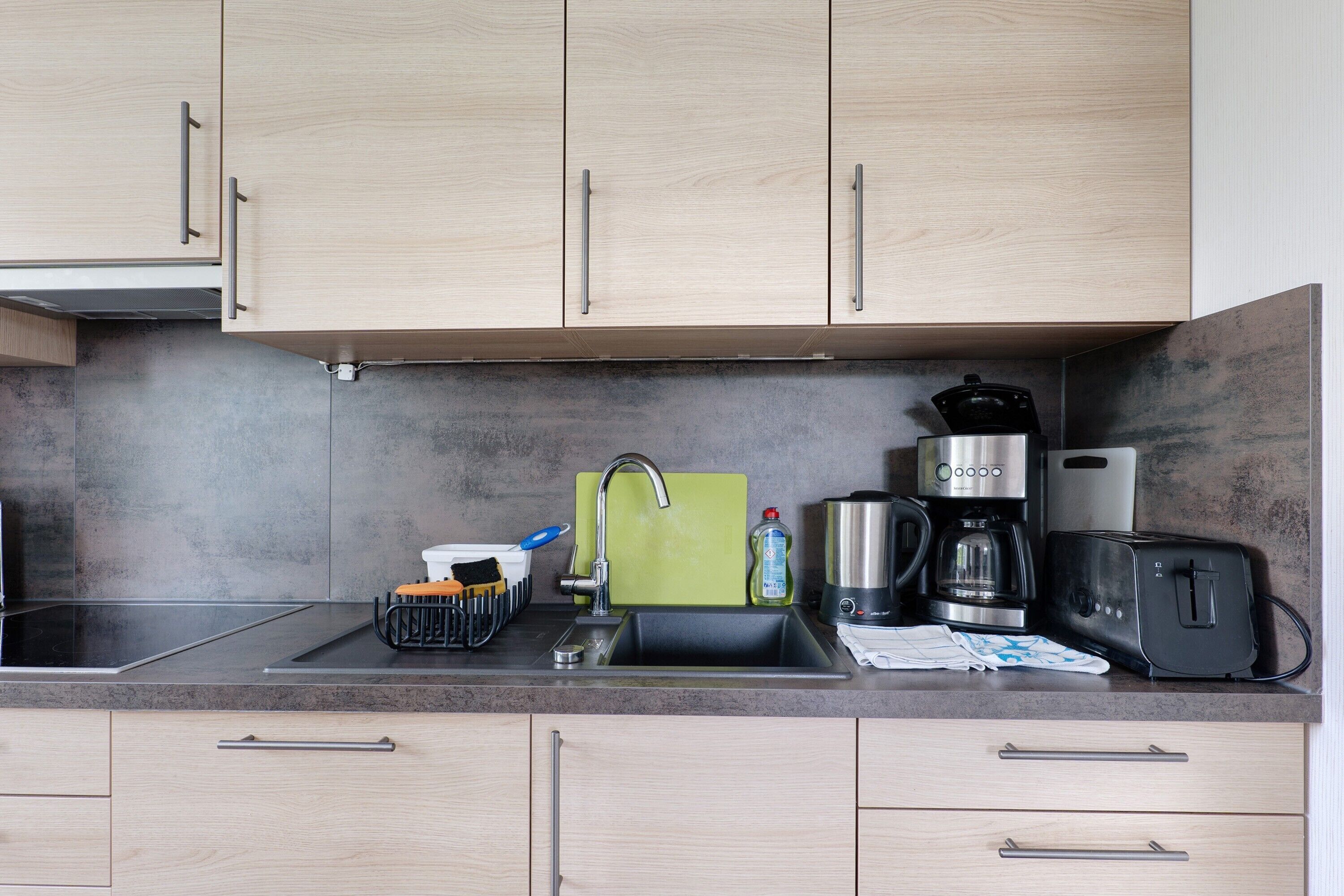 A modern kitchen with light wood cabinets, a black countertop, a sink, a coffee maker, a toaster, and a green cutting board.