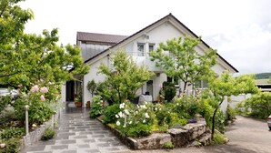 A two-story house with a paved driveway, a small garden with various plants and flowers, and a car parked in the driveway. - Holiday Apartment 'Trollinger' with Kitchenette, Balcony, and Wi-Fi (Vaihingen an der Enz)