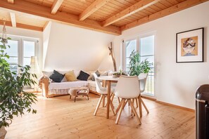 Ein gemütliches Wohnzimmer mit Holzboden, einem weißen Sofa mit Kissen, einem Esstisch mit Stühlen und einem Fenster mit Aussicht.