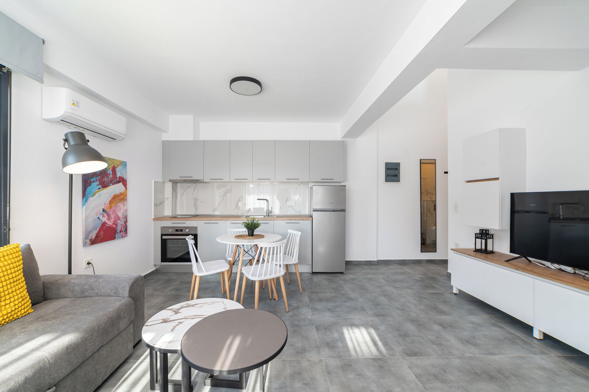 Een moderne keuken met witte kasten, een inbouwoven en een koelkast. Er is een eethoek met een tafel en stoelen, en een grijze bank met een geel kussen.