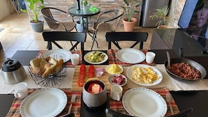 A dining table set with plates, bowls, and cutlery, surrounded by chairs, with potted plants and a laptop in the background. - JKIA Boutique Inn (Syokimau)