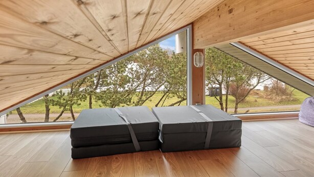 Ein Zimmer mit schrägen Wänden, Holzbalken und einem großen Fenster mit Blick auf Bäume und einen Zaun. Auf dem Boden liegen zwei schwarze Kissen.