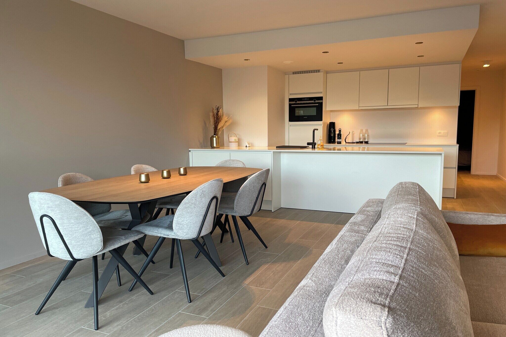 Une cuisine moderne avec des meubles blancs, un îlot central et un coin repas comprenant une table en bois et des chaises grises.