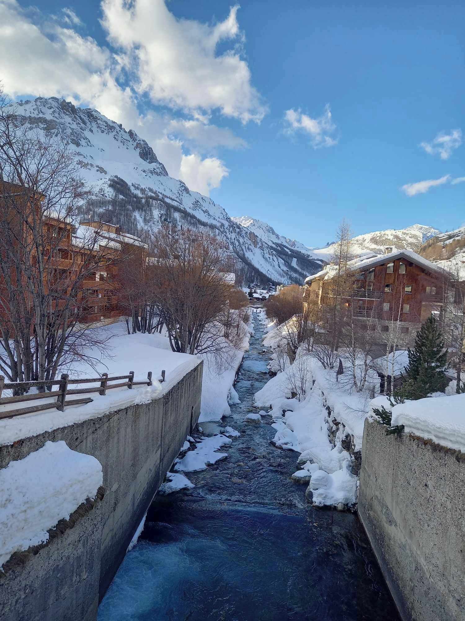 A snow-covered mountain landscape with a river flowing through a concrete channel, surrounded by wooden buildings and bare trees.