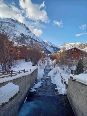 Paisaje montañoso cubierto de nieve con un río que fluye por un canal de hormigón, rodeado de edificios de madera y árboles sin hojas.