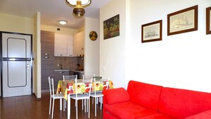 Ein Wohnzimmer mit rotem Sofa, eine Essecke mit Tisch und Stühlen sowie eine Küche mit Schränken und einem Kühlschrank.