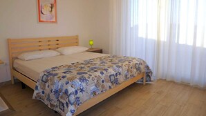 Een slaapkamer met een houten bed, een nachtkastje, een lamp en een raam met dunne gordijnen.