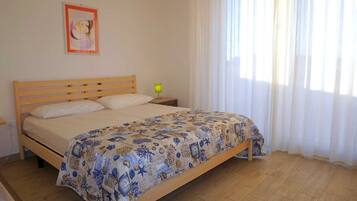 A bedroom with a wooden bed, a nightstand, a lamp, and a window with sheer curtains.