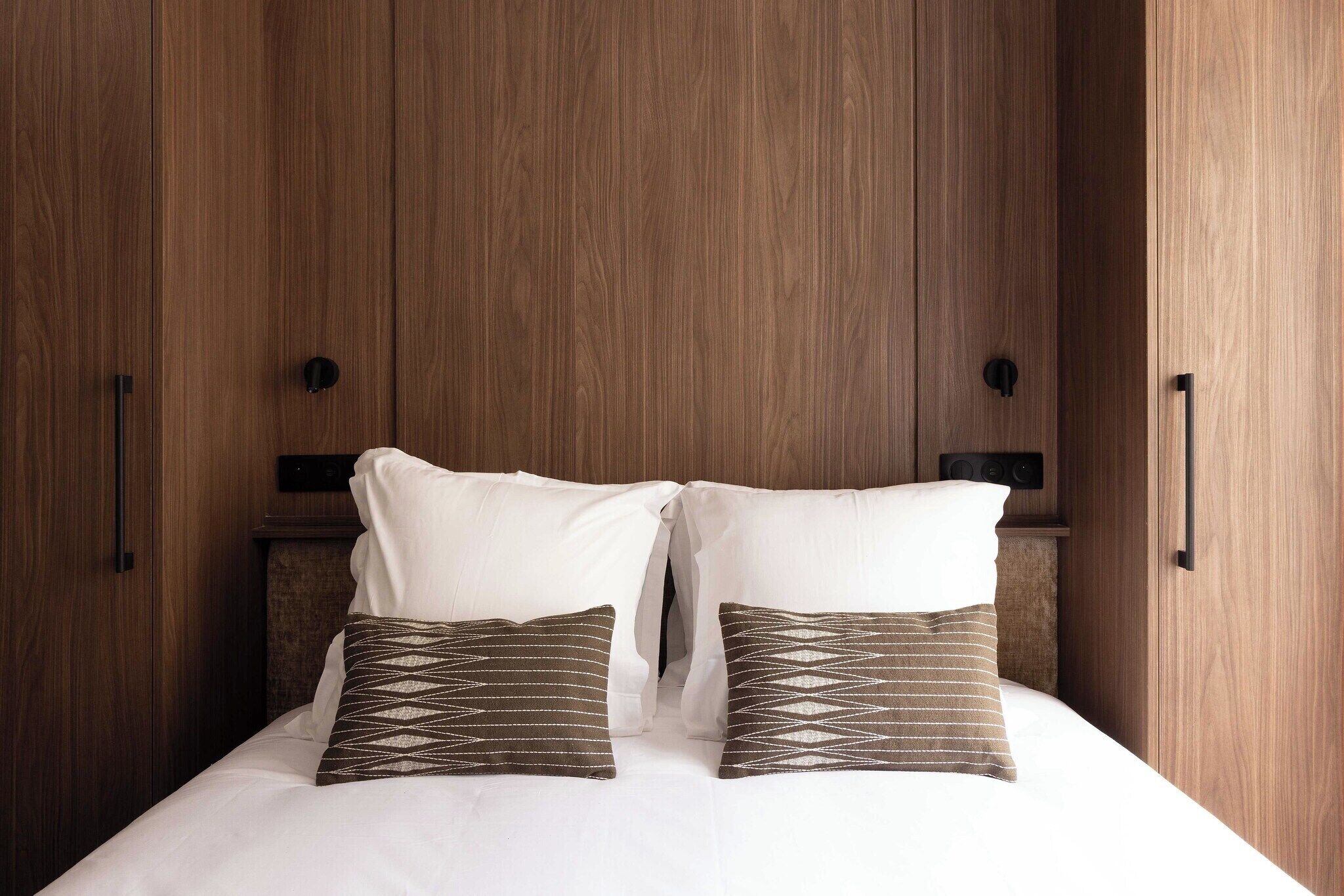 A bedroom with a wooden wardrobe, a bed with white bedding, and two patterned pillows.