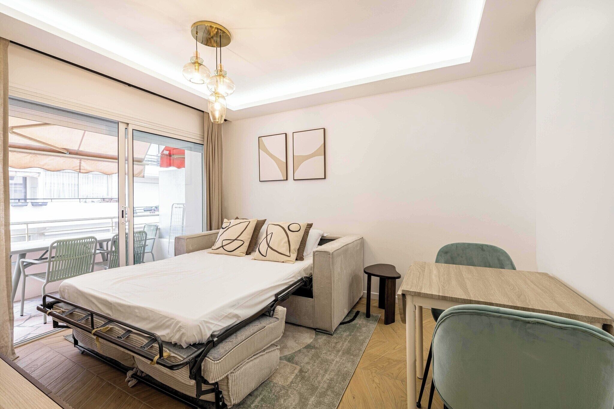 A compact hotel room with a bed, a desk, and a chair. There is a sliding door leading to an outdoor area with chairs.