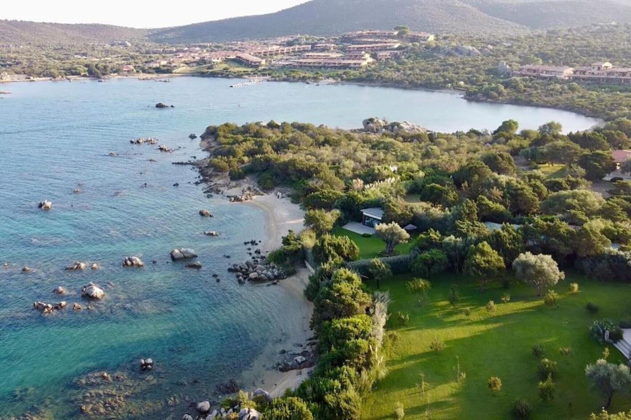 An aerial view of a coastal area with a beach, greenery, and buildings in the background.