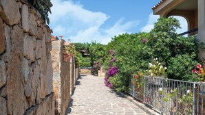 A stone-paved alleyway flanked by a stone wall and a metal fence with flowering plants. - Apartment in Marinella near the beach (Marinella)