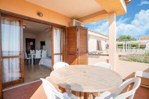 A balcony with a wooden table and chairs, a sliding glass door leading to a room with a TV, and a view of the surrounding area. - Apartment in Sardinia near stunning beaches (Marinella)