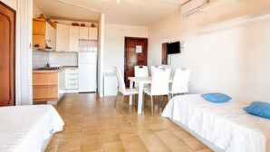 A room with two beds, a dining table with chairs, a kitchen area, and a TV mounted on the wall. - Apartment in Sardinia near stunning beaches (Marinella)