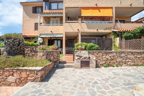 A multi-story building with a stone facade, balconies, and a paved entrance area with stone planters. - Apartment in Sardinia near stunning beaches (Marinella)