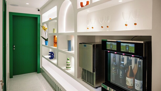 A hotel lobby with a green door, a wine cooler displaying various bottles, and shelves with decorative items. - Pepella (Piano di Sorrento)