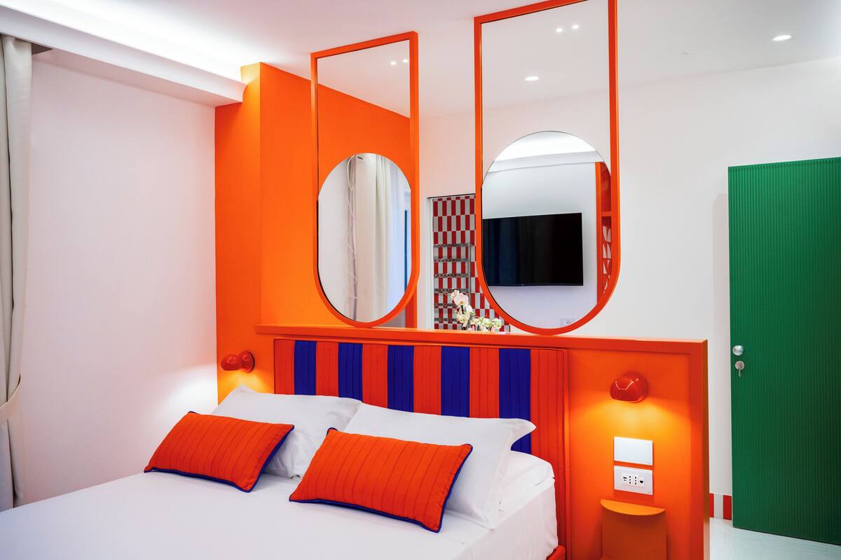 Een moderne hotelkamer met een bed, oranje accenten, een spiegel en een groene deur.