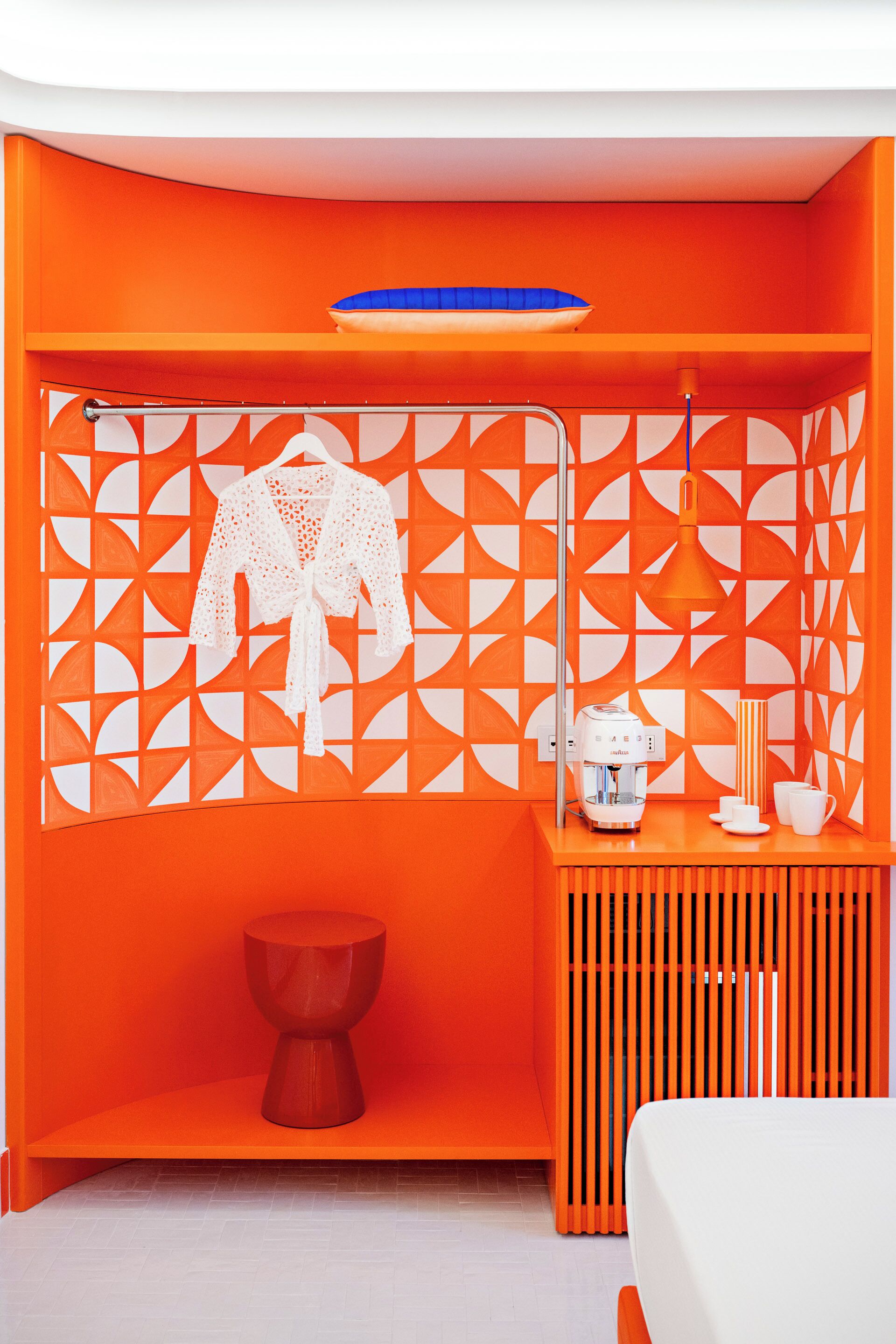A room with a vibrant orange color scheme, a white bed, a wooden side table with a coffee maker, cups, and a lamp.
