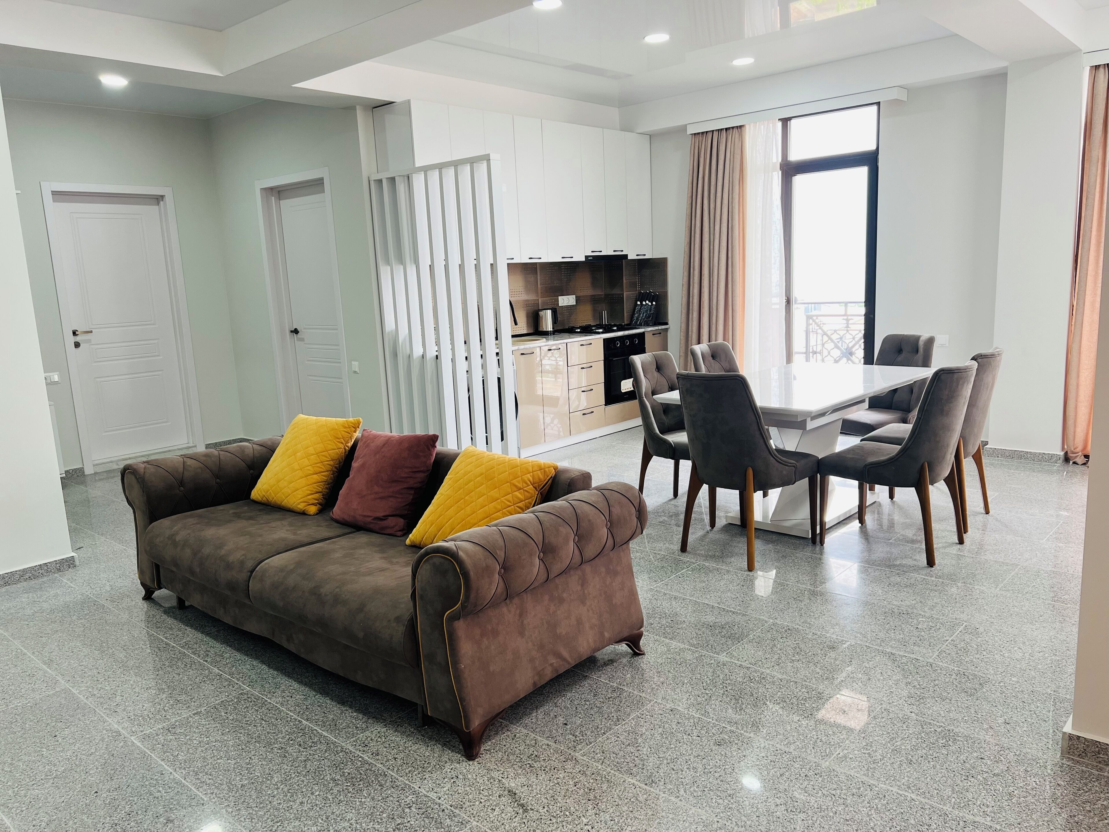 A modern living room with a brown sofa, dining table, and chairs, a kitchen area, and a large window with curtains.