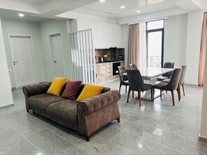 A modern living room with a brown sofa, dining table, and chairs, a kitchen area, and a large window with curtains. - Panorama David (Tbilisi)
