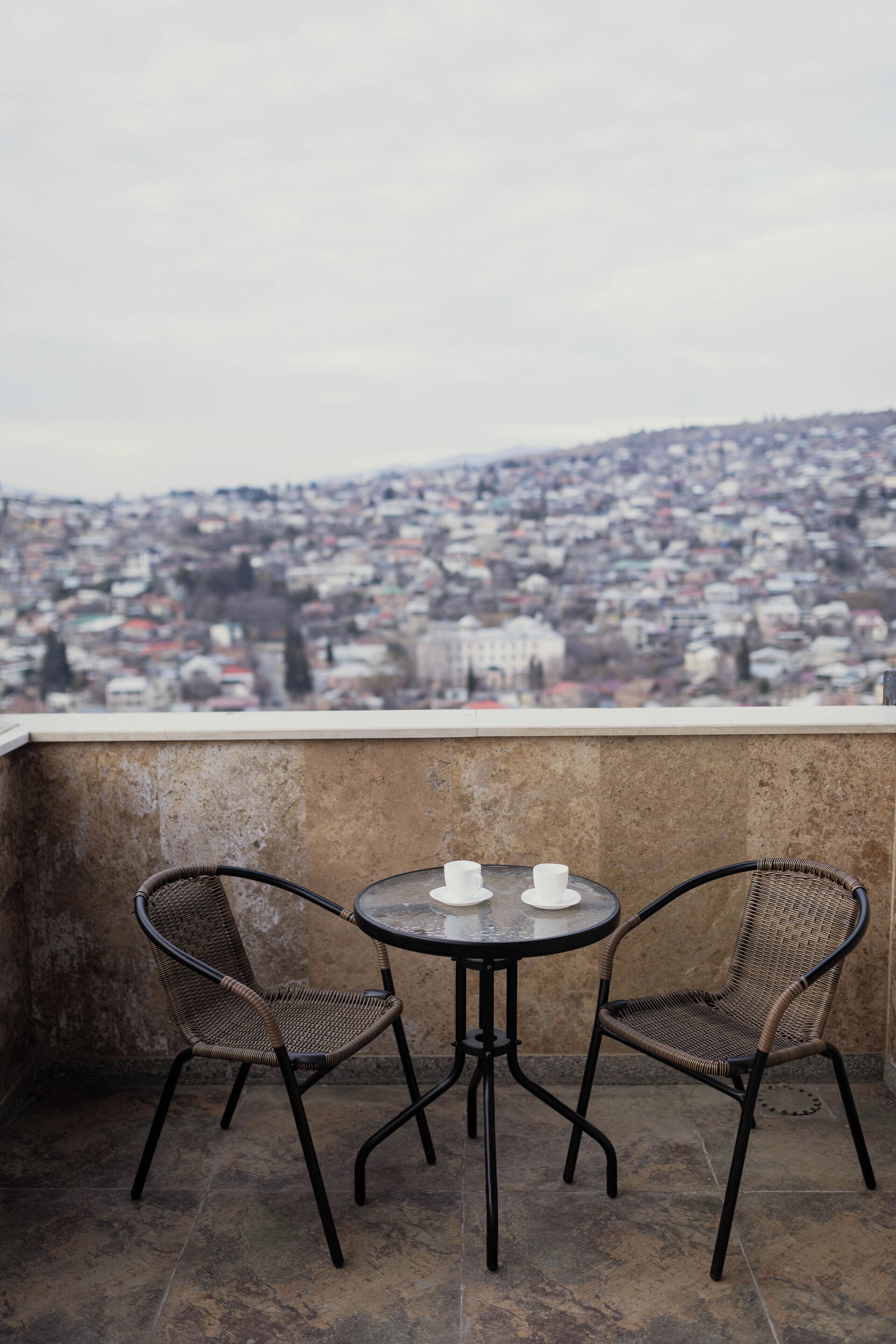 A balcony with two wicker chairs and a small round table, overlooking a densely packed cityscape.
