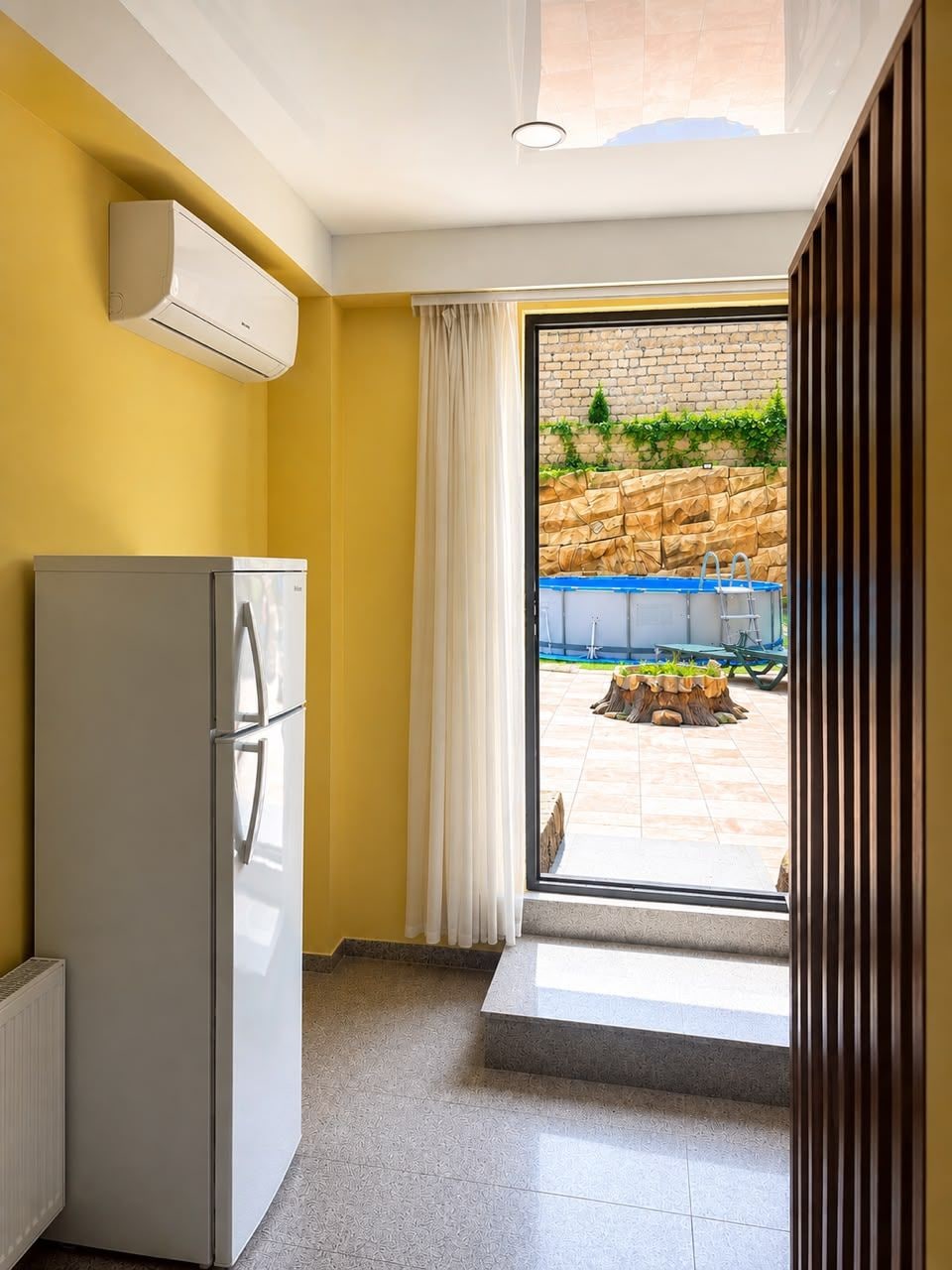 Cuina compacta amb nevera integrada, finestra que dona a zona exterior amb piscina i mur de pedra.