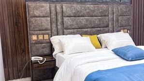 A modern bedroom with a grey stone wall, a wooden panel on one side, a grey upholstered headboard, a white bed with blue and yellow pillows, and a small bedside table with a white phone. - Dreamland suites (Abuja)