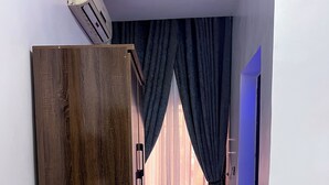 A hotel room with a wooden wardrobe, a white cabinet, a bed, a bathroom with a mirror, and a view of the outside through a window with blue curtains. - Dreamland suites (Abuja)