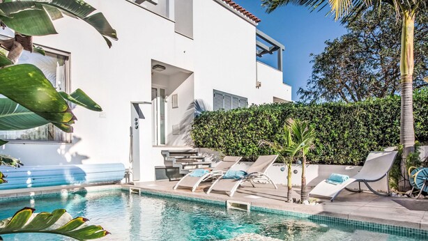 Un área de piscina moderna con agua azul cristalina, sillas de descanso y un edificio blanco con balcón.