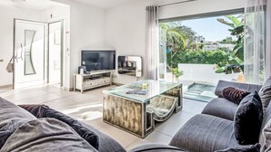 Una sala moderna con una mesa de centro de vidrio, un sofá, un televisor y un ventanal corredero que da a un balcón con vistas a zonas verdes y edificios.