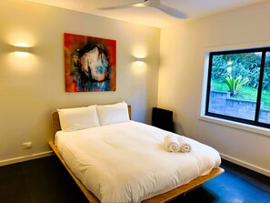 A bedroom with a bed, a painting, a window with a view of greenery, and a ceiling fan.