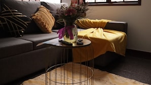 Un salon avec un canapé, une petite table sur laquelle est posé un vase de fleurs et une couverture jaune sur l’accoudoir.