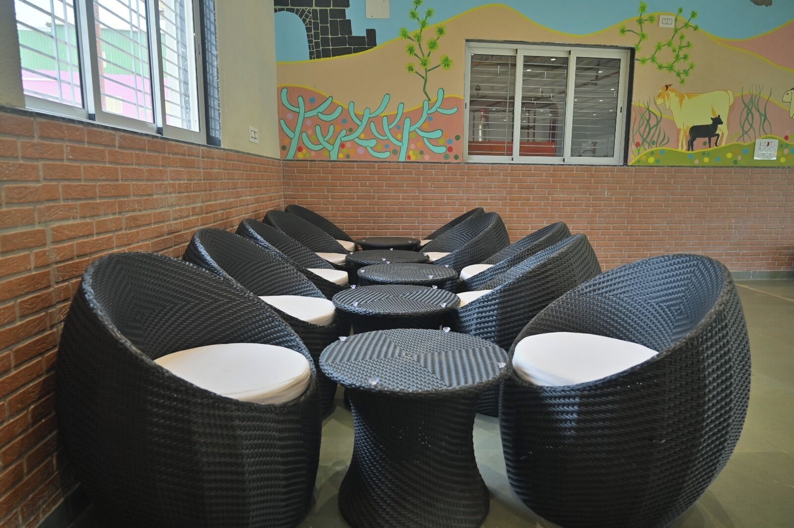 A row of wicker chairs with white cushions arranged around a central table in a room with brick walls and a mural depicting a landscape with trees and animals.