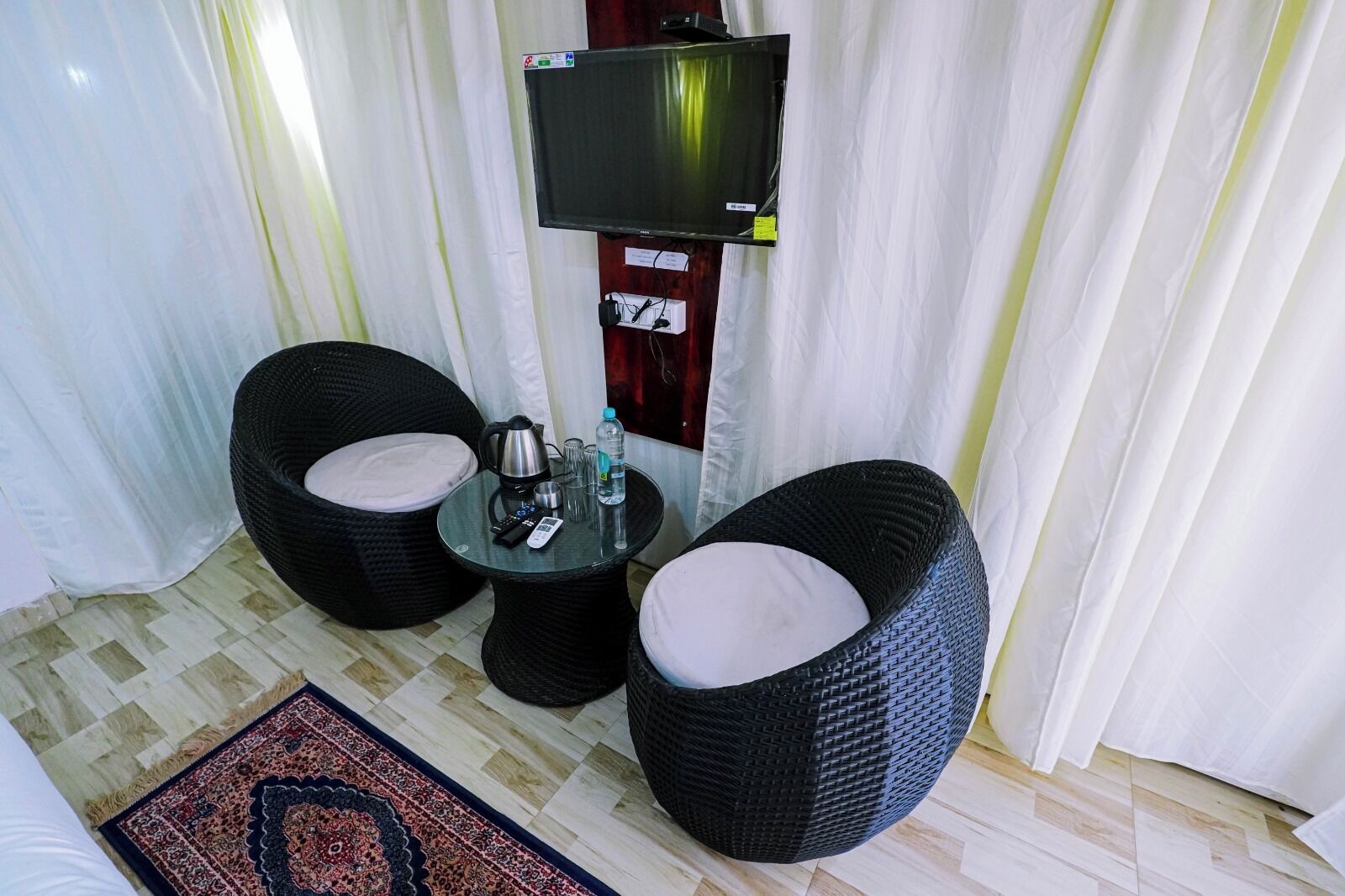 A room with two wicker chairs, a small table with a kettle, cups, and a bottle, and a TV mounted on the wall.