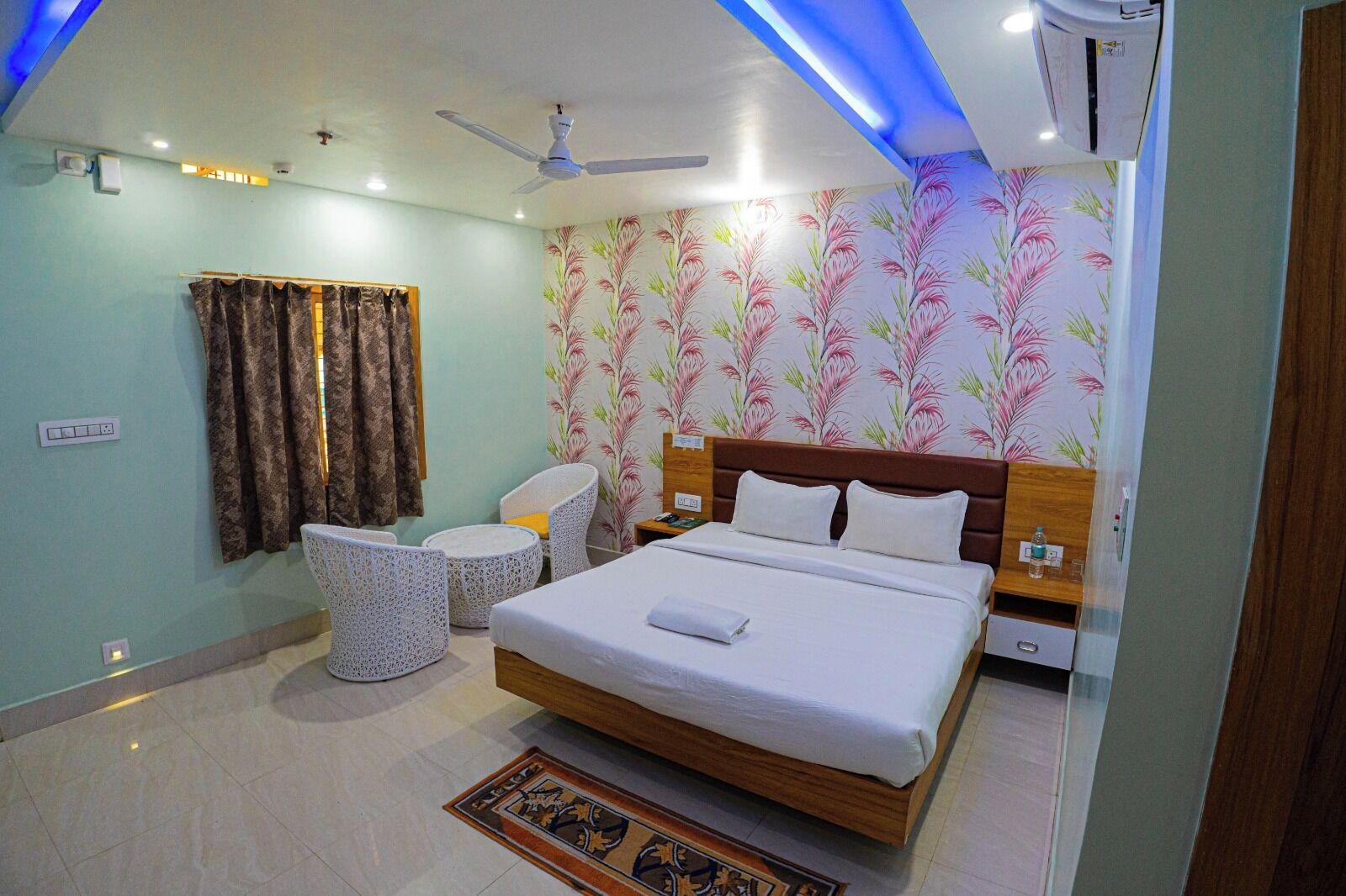 A hotel room with a bed, two chairs, a small table, a ceiling fan, and a patterned wall.
