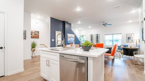 A modern kitchen with white cabinetry, a central island, and a dining area with orange chairs. - Game Day Home Base | Steps to Globe Life & AT&T (Arlington)