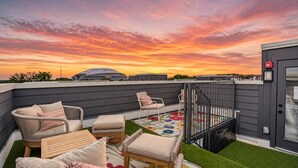 A rooftop patio with wicker furniture, a colorful rug, and a view of a stadium at sunset. - Game Day Home Base | Steps to Globe Life & AT&T (Arlington)