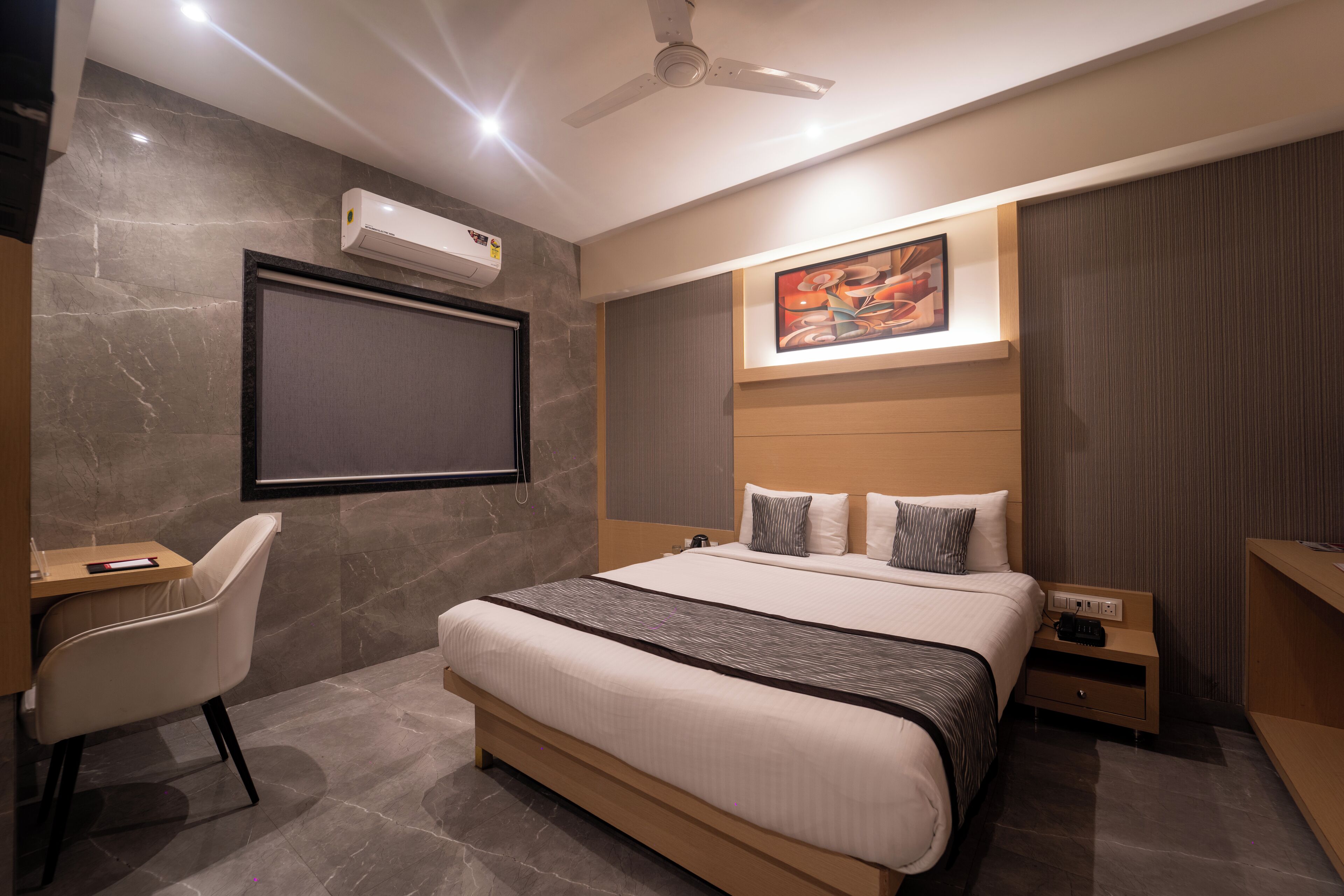 A modern hotel room with a large bed, a desk, a chair, a ceiling fan, an air conditioner, and a painting on the wall.