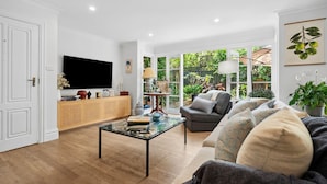 A spacious living room with a large flat-screen TV, a glass coffee table, a grey sofa, and a wooden cabinet. - Reed Cottage - Large private garden cottage a perfect base for your Sydney stay (Cremorne)