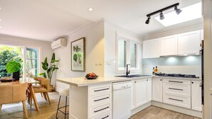 A modern kitchen with white cabinetry, a dishwasher, and a gas stove. There is a dining area with a wooden table and chairs, and a large window with plants outside. - Reed Cottage - Large private garden cottage a perfect base for your Sydney stay (Cremorne)
