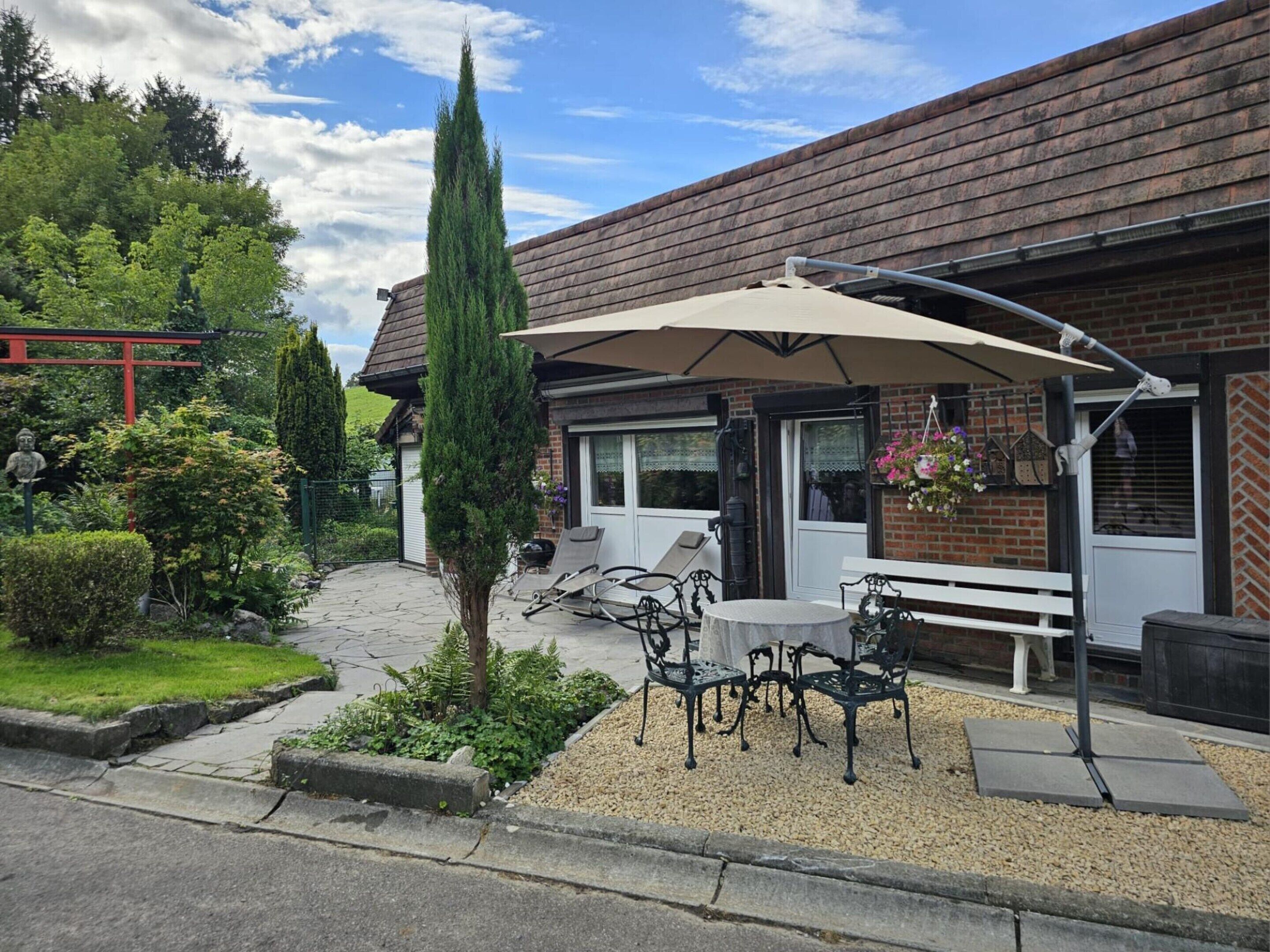 A patio area with outdoor seating, a tall conifer tree, and a brick building with a tiled roof.