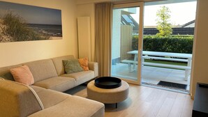 A modern living room with a beige sofa, a round ottoman, and a glass door leading to an outdoor patio with a white table and benches. - Apartment Near Marina Nieuwpoort & Pool (Nieuwpoort)