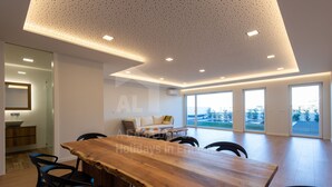 Una habitación amplia con una gran mesa de madera y sillas, un sofá, y vistas al exterior a través de amplios ventanales.